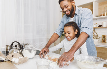 Happy afro man and his child daughter rolling up dough together, having fun. Home bakery concept,...
