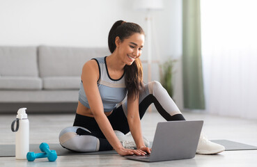 Smiling beautiful lady in sportswear getting ready for online fitness class from home, sitting on...