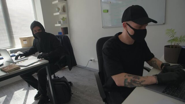 Handheld shot of masked man hacking into laptop in office then passing folder with confidential documents to accomplice stealing secret data in hurry during industrial espionage