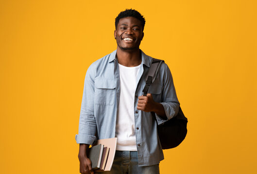 Positive millennial black man student in casual posing with books and notepads on yellow studio background, young african american guy enjoying studying at university or college, copy space