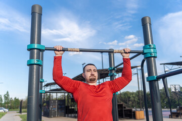 Man in red shirt doing pull-ups at an outdoor gym. The blue sky suggests a sunny day for a workout and active lifestyle