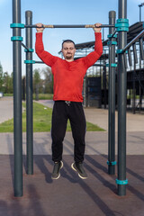 Man in red shirt doing pull-ups at an outdoor gym. The blue sky suggests a sunny day for a workout and active lifestyle
