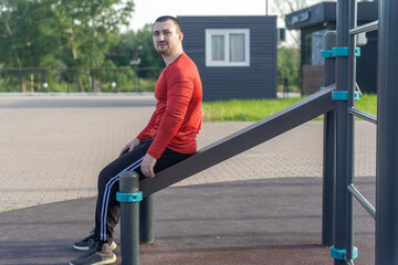 Man does sit-ups on a bench at an outdoor gym. Red shirt, black pants, showcasing a workout routine for healthy lifestyle. Blue sky background at a park.