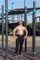 Muscular man doing pull-ups on outdoor gym equipment. Shirtless, he showcases defined upper body strength. Set in a sunny park, the photo captures a healthy lifestyle
