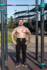 Muscular man doing pull-ups on outdoor gym equipment. Shirtless, he showcases defined upper body strength. Set in a sunny park, the photo captures a healthy lifestyle