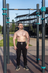 Muscular man doing pull-ups on outdoor gym equipment. Shirtless, he showcases defined upper body strength. Set in a sunny park, the photo captures a healthy lifestyle