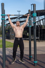 Muscular man doing pull-ups on outdoor gym equipment. Shirtless, he showcases defined upper body strength. Set in a sunny park, the photo captures a healthy lifestyle