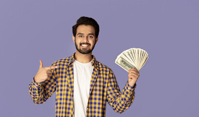 Smiling Indian man pointing at fan of money over lilac background, copy space