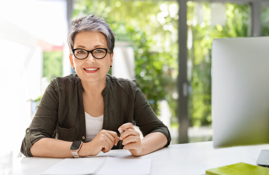 Cheerful attractive mature woman in smart casual wearing eyeglasses business consultant working at green eco-friendly office, sitting at workdesk in front of computer, holding pen, smiling at camera