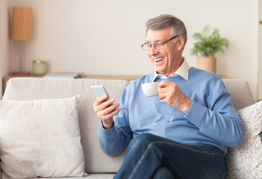 Happy Senior Gentleman Using Smartphone Texting And Having Coffee Sitting On Sofa At Home. Selective Focus