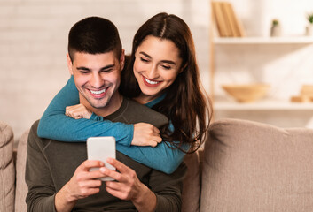 Romantic millennial couple sharing mobile sitting on couch at their apartment, girl hugging her man...