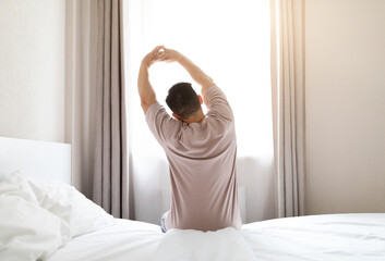 Back view of brunette man wearing pajamas sitting on bed and stretching body after waking up in the...