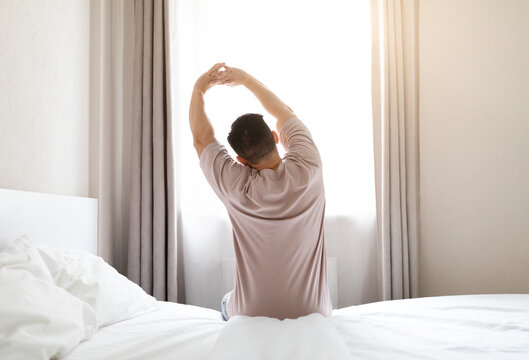 Back view of brunette man wearing pajamas sitting on bed and stretching body after waking up in the morning, looking at window, copy space. Comfortable healthy sleep concept - Powered by Adobe