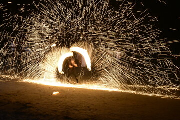 Man spinning burning steel wool at night, creating dramatic fire sparks on a beach.