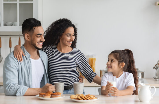 Happy arabic family having breakfast while spending weekend together at home, cheerful young father, mother and curly little girl sitting at kitchen, drinking coffee and milk with homemade cookies - Powered by Adobe
