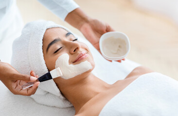 Anti-Aging Skincare. Cosmetologist applying clay mask on face of young indian woman, beautiful eastern lady with towel on head lying on table, enjoying beauty treatments in spa salon, closeup