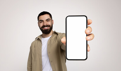 Happy millennial arab man with a beard, wearing a khaki shirt and white t-shirt, extends his arm to present a smartphone with a blank screen, inviting viewer interaction