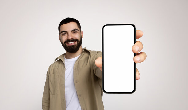 Happy millennial arab man with a beard, wearing a khaki shirt and white t-shirt, extends his arm to present a smartphone with a blank screen, inviting viewer interaction