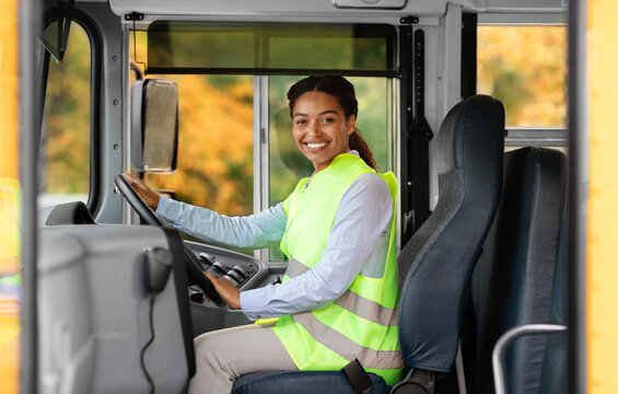 Happy black female driver sitting inside of bus and looking at camera, smiling beautiful african american woman in reflecting clothing holding steering wheel, waiting passengers, enjoying her work