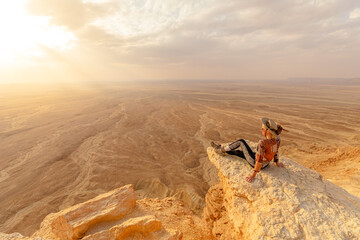 Female tourist wearing hat and sunglasses sitting on rock admiring sunset view of Tuwaiq...
