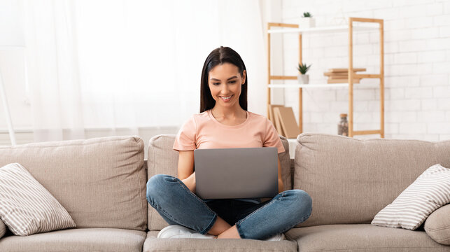 Frontal portrait of happy girl chatting online on laptop with friend at home, sitting on beige couch in light living room - Powered by Adobe