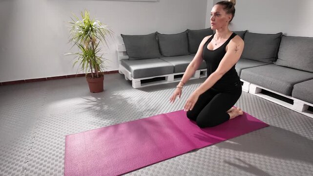 Woman performing Ustrasana (Camel Pose) yoga posture on a pink mat at home. She is dressed in black sportswear, kneeling and leaning backward while holding her heels. Practicing in a calm, modern indo