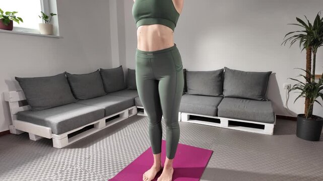 Rear view of a woman performing Uttanasana (standing forward bend) yoga pose at home. She is wearing a green sports outfit, practicing on a pink yoga mat in a modern, minimalistic indoor setting with 