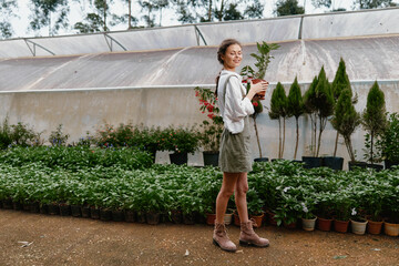Young woman in a greenhouse holding plants, wearing a white blouse and green skirt, enjoying nature...