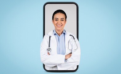 Fototapeta premium Young brazilian woman doctor in white uniform posing with stethoscope and folded arms in huge cellphone screen, blue background, banner. Medical services concept