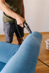Vertical cropped shot of cleaning service employee sanitizing blue sofa with high-temperature steam cleaner in contemporary living space, removing dirt and bacteria. Concept of hygiene and cleanliness