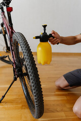 Close-up of cyclist using yellow handheld water sprayer to clean dusty dirty tire wheel of mountain bike at home. Concept of bike maintenance, service and cleaning practices
