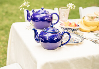 two glossy blue ceramic teapots arranged on a white tablecloth at an outdoor garden table, with floral tableware, fruit bowl, and fresh bananas in a bright picnic