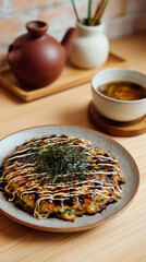 A close up of okonomiyaki on a plate with a tea pot and soup on a wooden table top setting