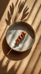 A skewer of grilled meat on a plate casting shadows on a wooden surface in the bright sunlight