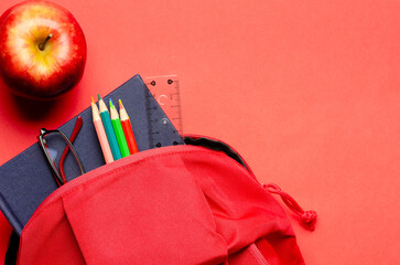 A red backpack with a notebook, a pair of glasses, a ruler and colorful pencils on red background and an apple. Back to school concept with copy space. 