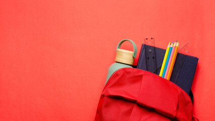 A red backpack with a notebook, a ruler, a square, colorful pencils and a bottle on red background. Back to school concept with copy space. 