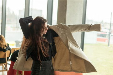 A woman is currently trying on a stylish coat in front of a large window, showcasing her...
