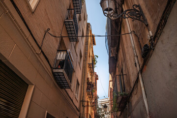 Narrow street in Ciutat Vella, Valencia, Spain, where brick and stucco walls, iron balconies, hanging cables and an old street lamp frame a slice of blue evening sky.