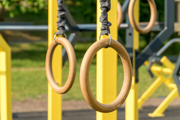 Two wooden gymnastic rings hang from a structure in a fitness park, surrounded by greenery,...
