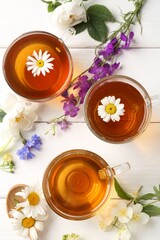 Aromatic herbal tea in glass cups, different flowers and green leaves on white wooden table, flat lay