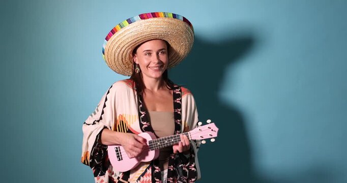 Happy woman in sombrero hat playing ukulele and singing on light blue background