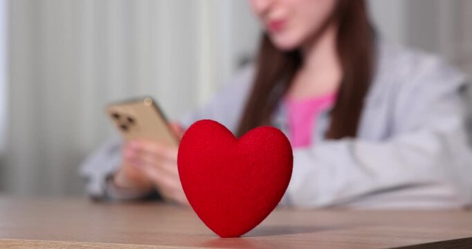 Online dating site. Woman using smartphone at table indoors, focus on decorative heart