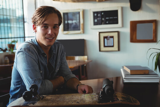 Man sitting at wooden desk near window, holding skateboard deck and browsing through stack of books