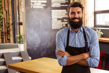 Adult male barista wearing black apron smiling in coffee shop showing chalkboard menu, copy space
