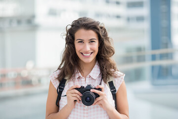Woman standing on rooftop balcony holding DSLR camera wearing sleeveless plaid shirt and backpack