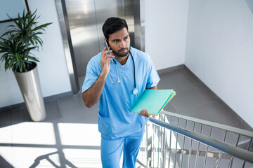 Asian male worker with stethoscope climbing hospital stairwell holding smartphone, colored folders