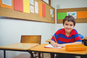 Primary schoolboy writing in notebook with yellow pencil at desk in classroom, copy space