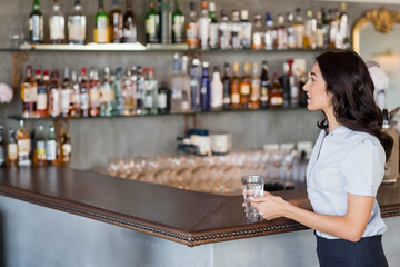 Woman standing at bar counter holding glass of water beside liquor bottles on shelves, copy space