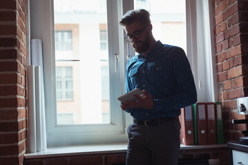 Bearded man leaning on sill in loft office reviewing tablet beside drawings and binders, copy space