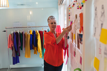 Senior female fashion designer pinning swatch to board in bright design studio with garment rack